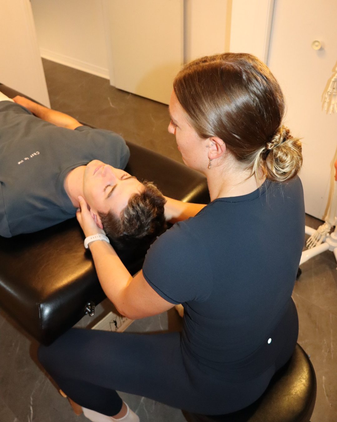 Maddie seated at the head of the treatment table, cradling a client’s head during craniosacral work.